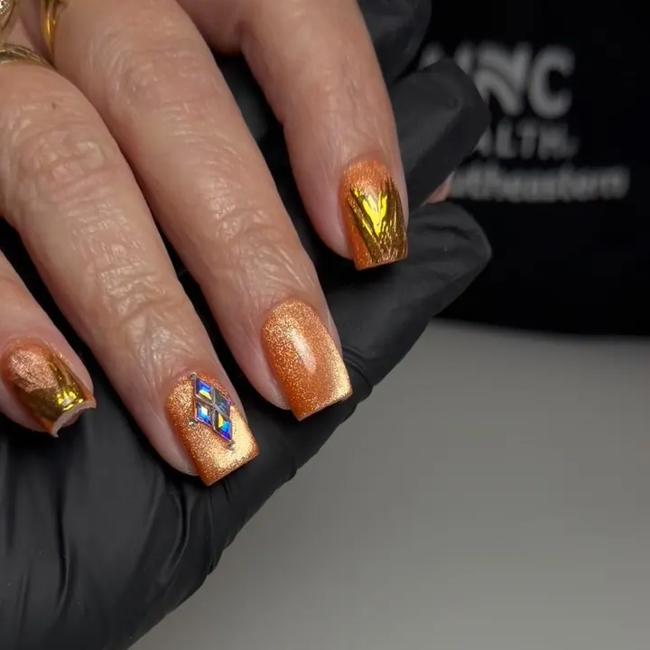 Uñas almendra cortas en cromo ámbar y dorado, con una uña de acento decorada con pequeños cristales.