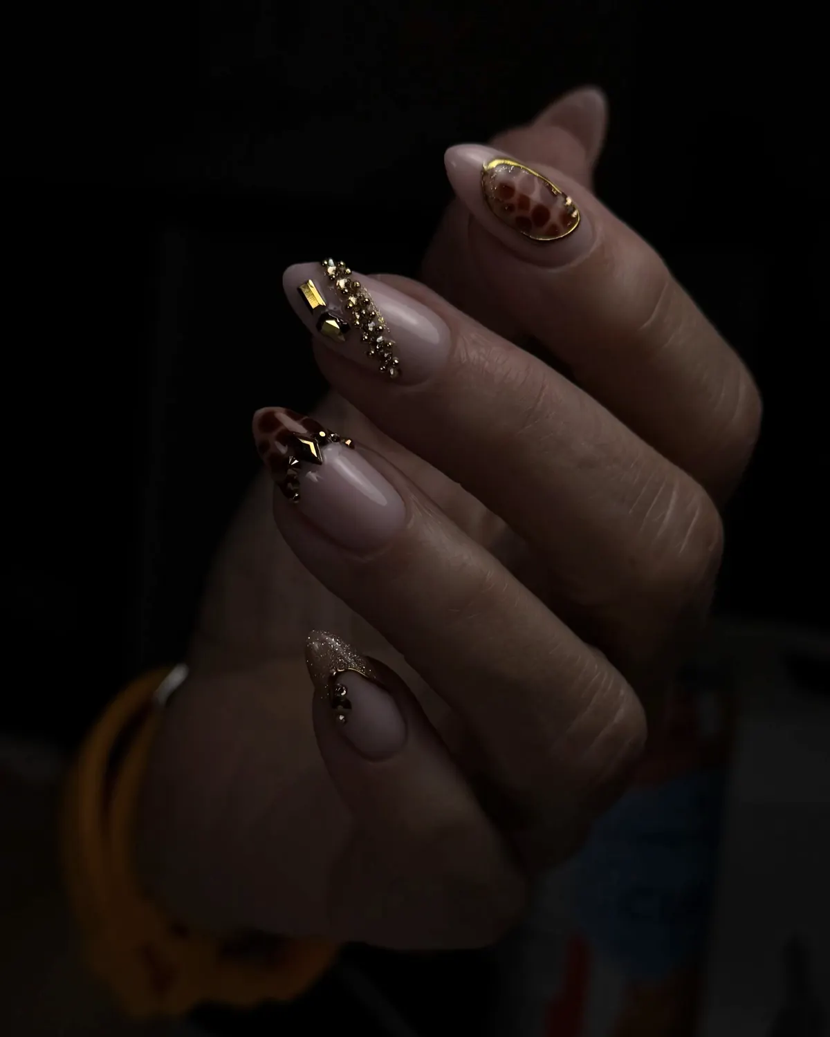 Uñas almendra esculpidas en tonos neutros con detalles de cristales, fotografiadas sobre fondo negro.