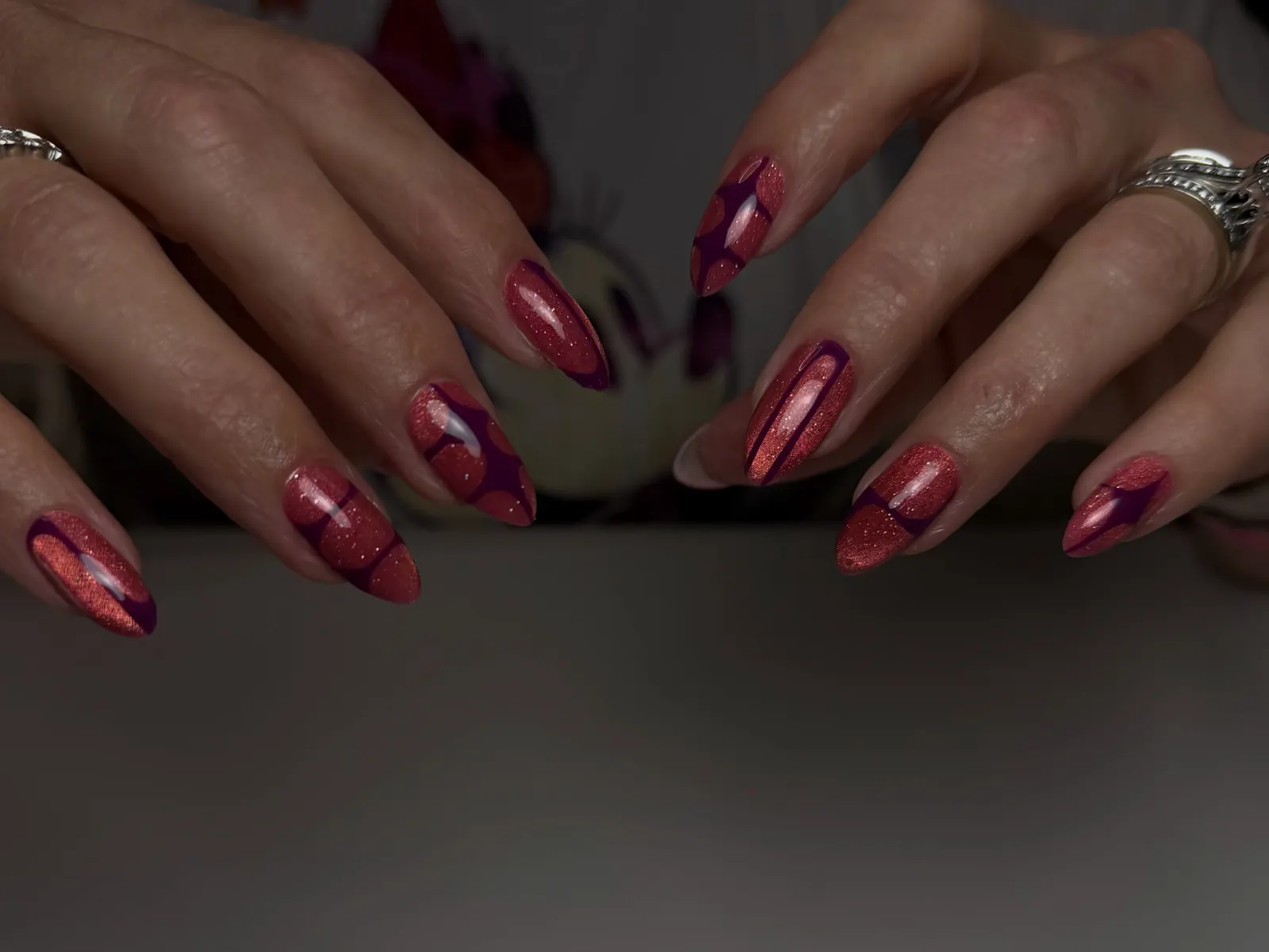 Uñas largas en punta en rosa cálido y burdeos con detalles metálicos, con los dedos abiertos sobre una mesa gris.