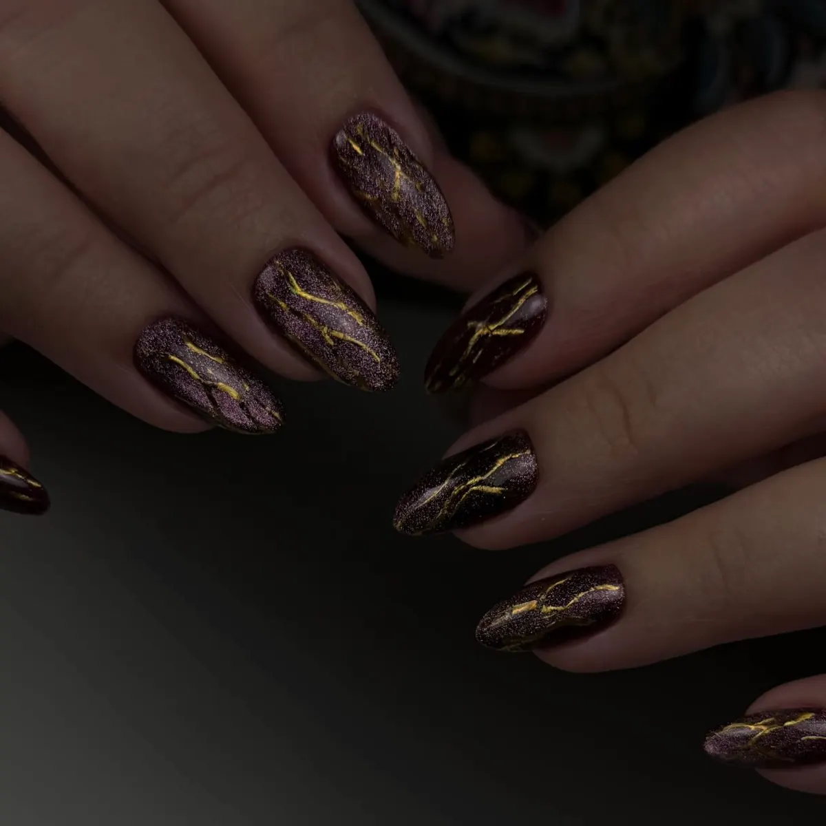 Uñas almendra esculpidas en metálico oscuro con finas líneas doradas, las manos sobre una mesa gris.