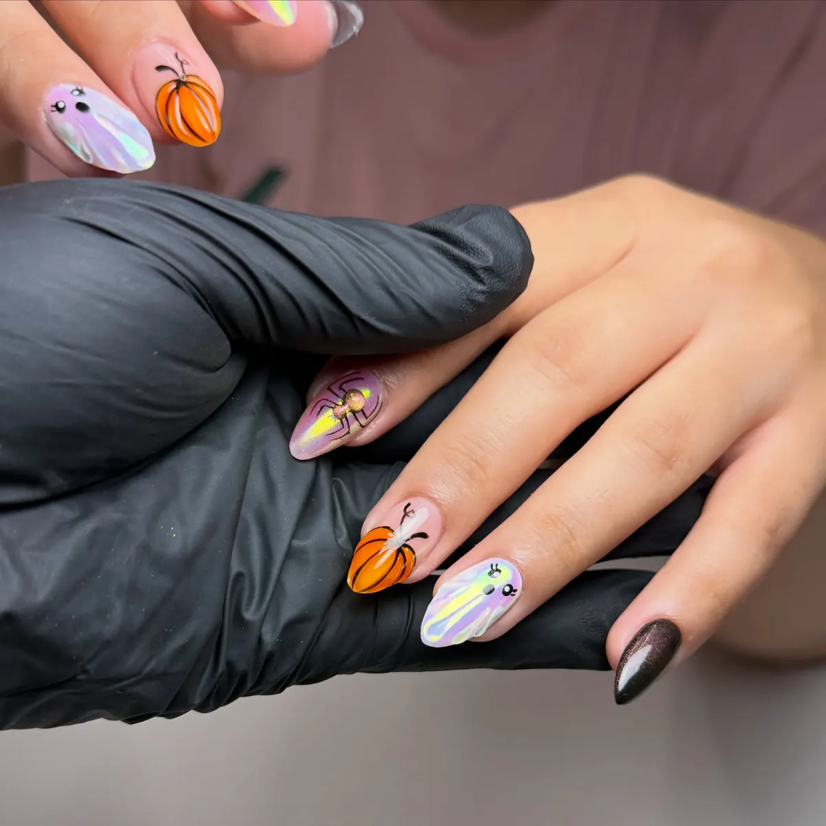 Arte de uñas con calabazas naranjas y pequeños fantasmas pintados sobre uñas iridiscentes, la mano apoyada sobre una mano con guante negro.
